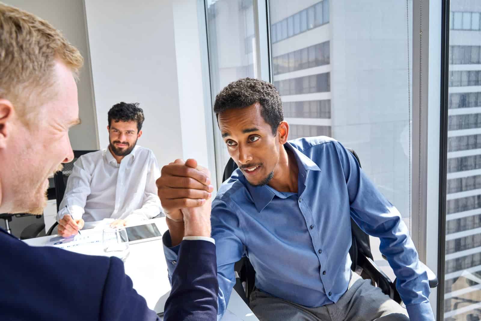 Diverse male coworkers opponents arm wrestling at group office team meeting.